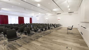 Een vergaderzaal met rijen zwarte stoelen, een houten vloer en witte muren.