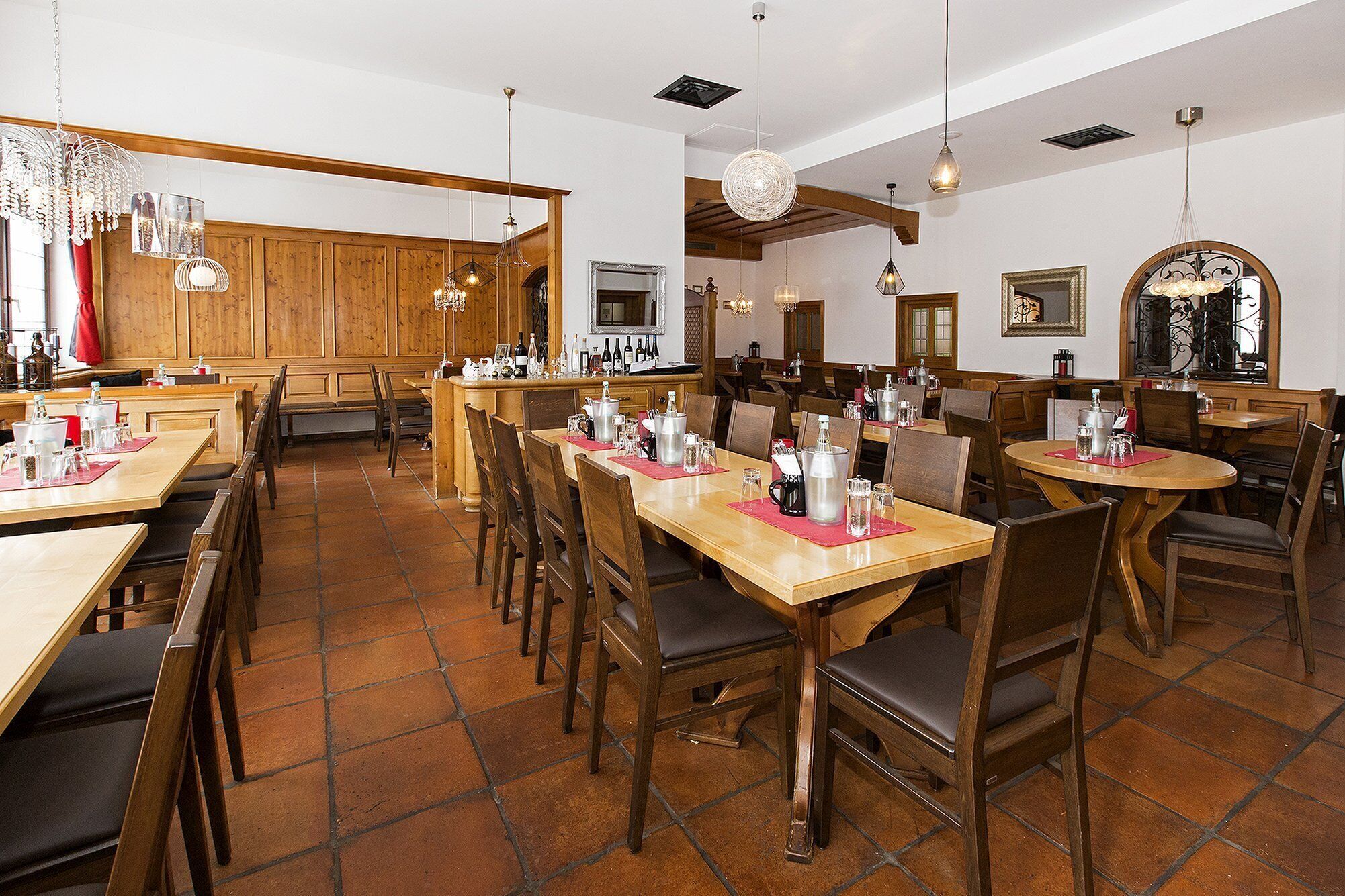 Un restaurante con mesas y sillas de madera, zona de bar y lámparas colgantes.