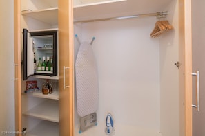 A closet with an open door revealing a shelf with bottles and glasses, an ironing board with an iron, and hangers on a rod. - Best Western Hotel Im Forum Muelheim (Muelheim an der Ruhr)