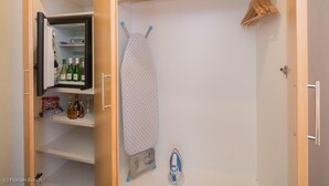 A closet with an open door revealing a shelf with bottles and glasses, an ironing board with an iron, and hangers on a rod. - Best Western Hotel Im Forum Muelheim (Muelheim an der Ruhr)