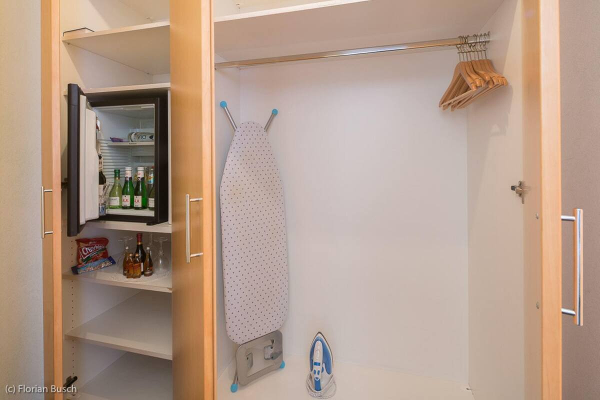 A closet with an open door revealing a shelf with bottles and glasses, an ironing board with an iron, and hangers on a rod.