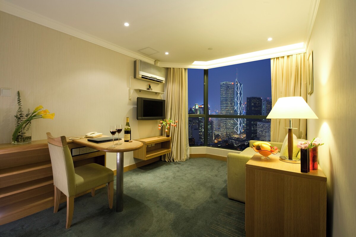 A hotel room with a dining area, a TV, a desk, a lamp, and a view of the cityscape at night.