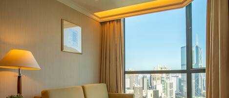 A hotel room with a large window, a sofa, a bedside table with a lamp, and a framed picture on the wall.