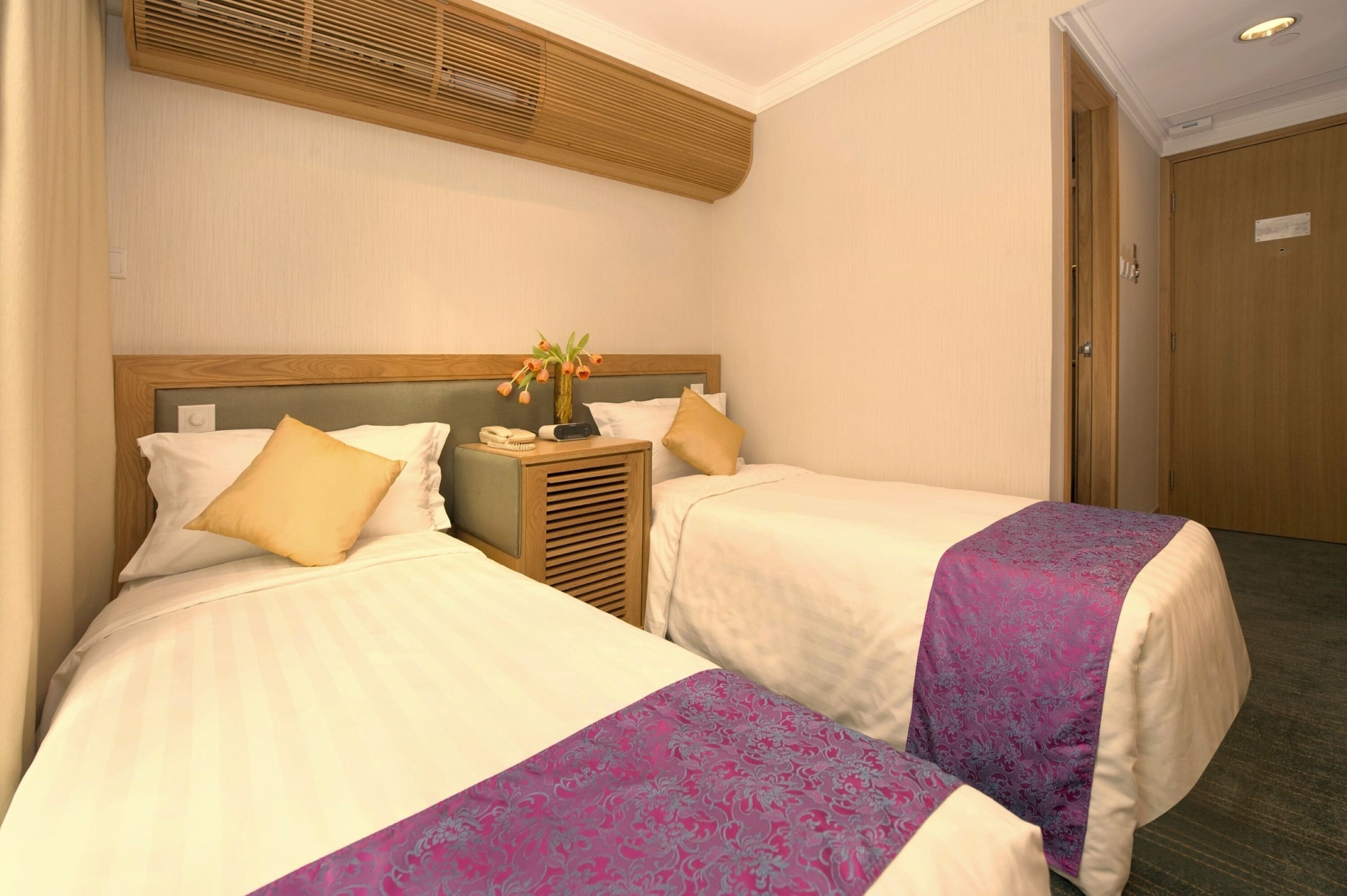 A hotel room with two beds, each with a wooden headboard, white linens, and a purple decorative runner. There is a nightstand with a lamp and a vase of flowers between the beds.