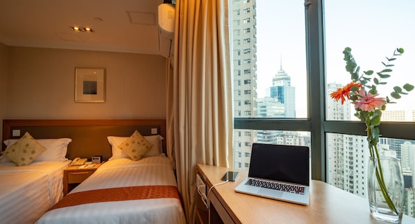 A hotel room with two beds, a desk with a laptop, a vase with flowers, and a view of a cityscape.