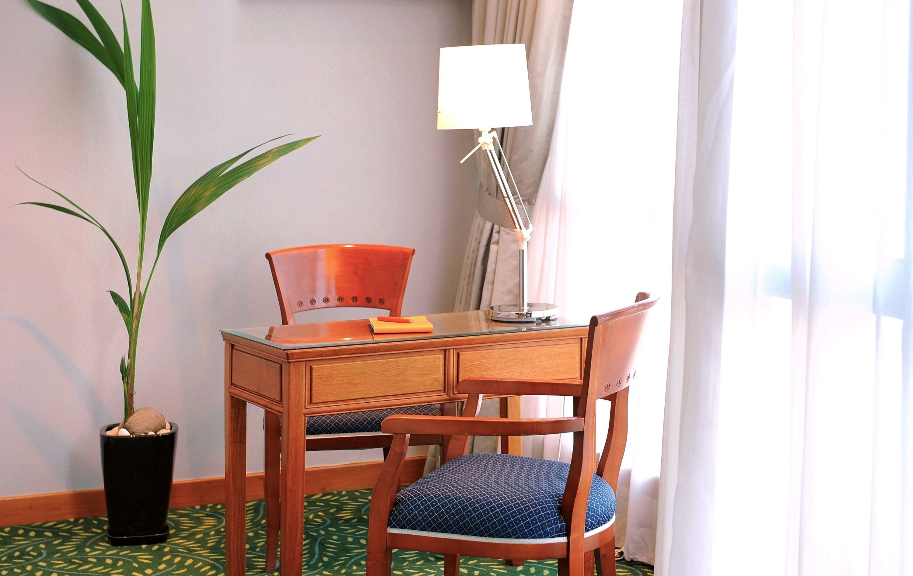 A hotel room with a wooden desk, a chair with a blue cushion, a potted plant, a lamp, and a window with curtains.