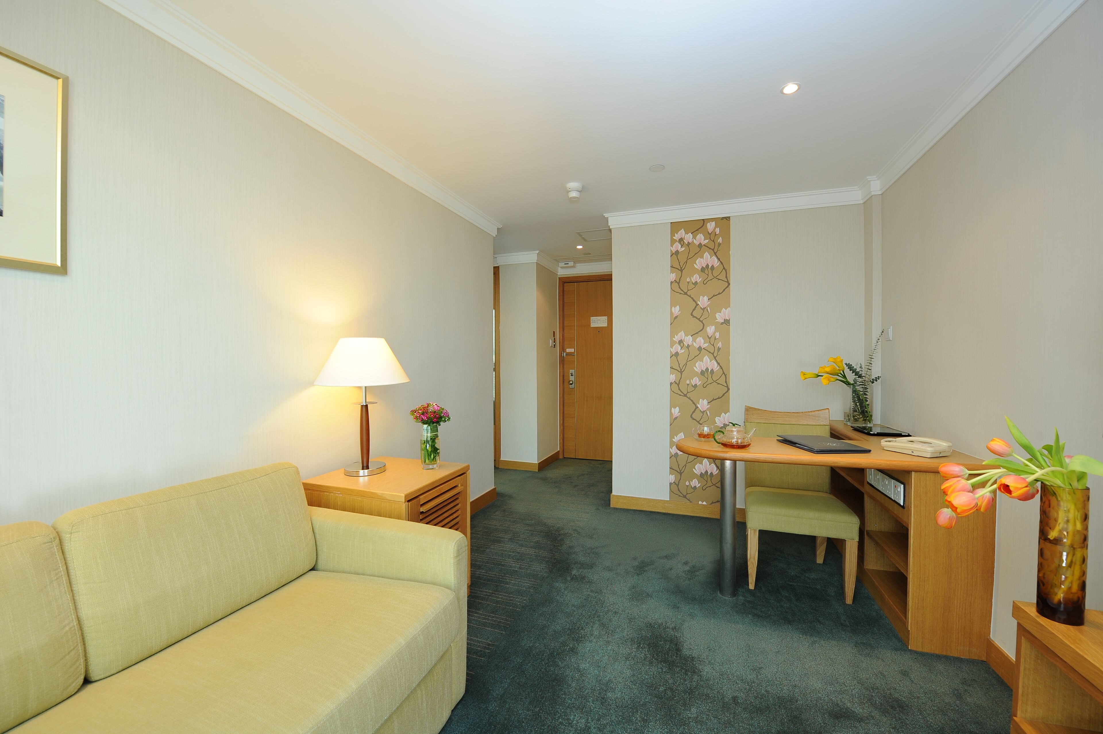 Habitación de hotel con sofá, escritorio, silla, lámpara y un jarrón con flores.