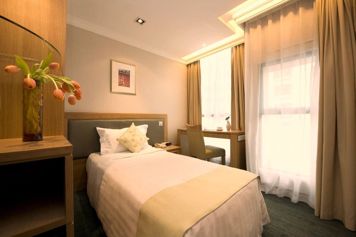 A hotel room with a bed, a desk, a chair, a vase with flowers, and a window with curtains.