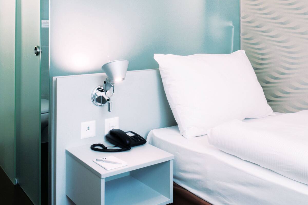 Habitación de hotel con cama, mesita de noche con lámpara, teléfono y almohada blanca.