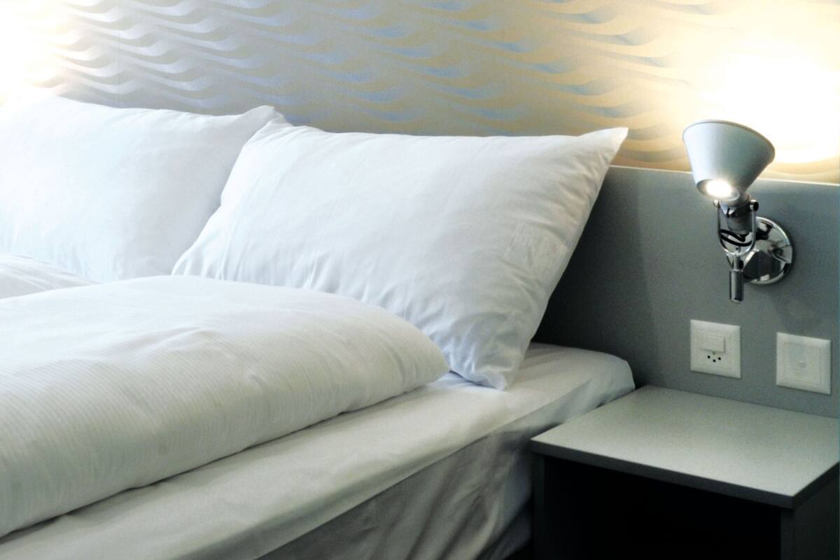 Una cama bien hecha con sábanas blancas, una lámpara de pared y un papel tapiz estampado.