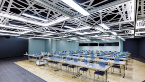 Un salón de conferencias con filas de mesas y sillas, techo alto con vigas a la vista y una pantalla grande al frente.