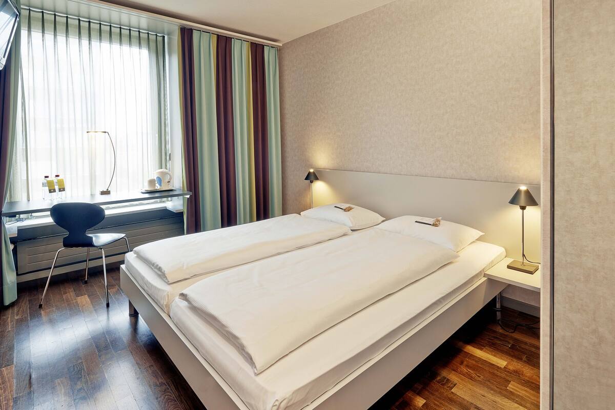 Una cama doble con ropa blanca, un escritorio con silla y una ventana con cortinas.