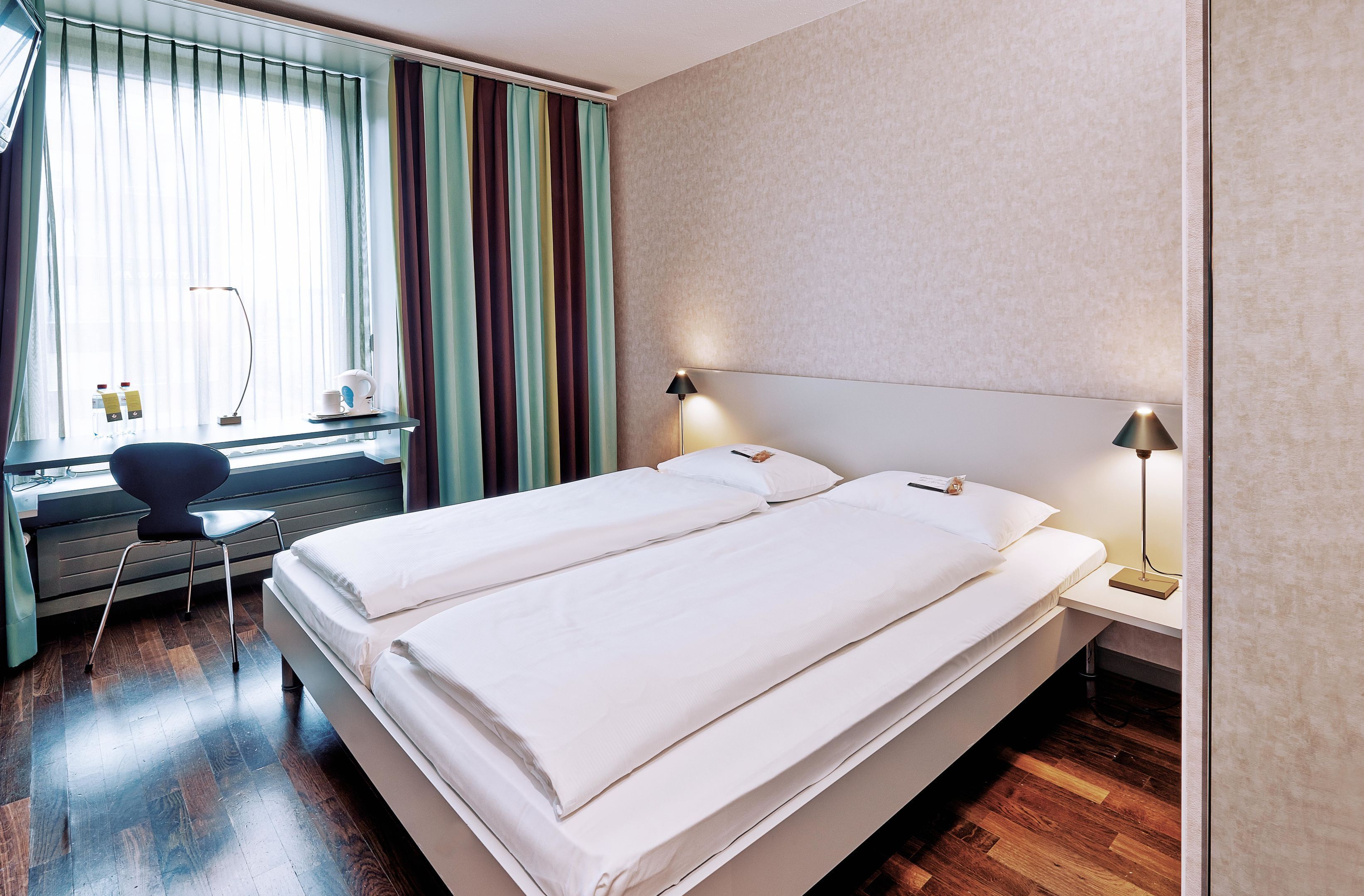 A double bed with white linens, a desk with a chair, a lamp, and a window with curtains.