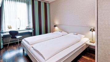 A double bed with white linens, a desk with a chair, a lamp, and a window with curtains.