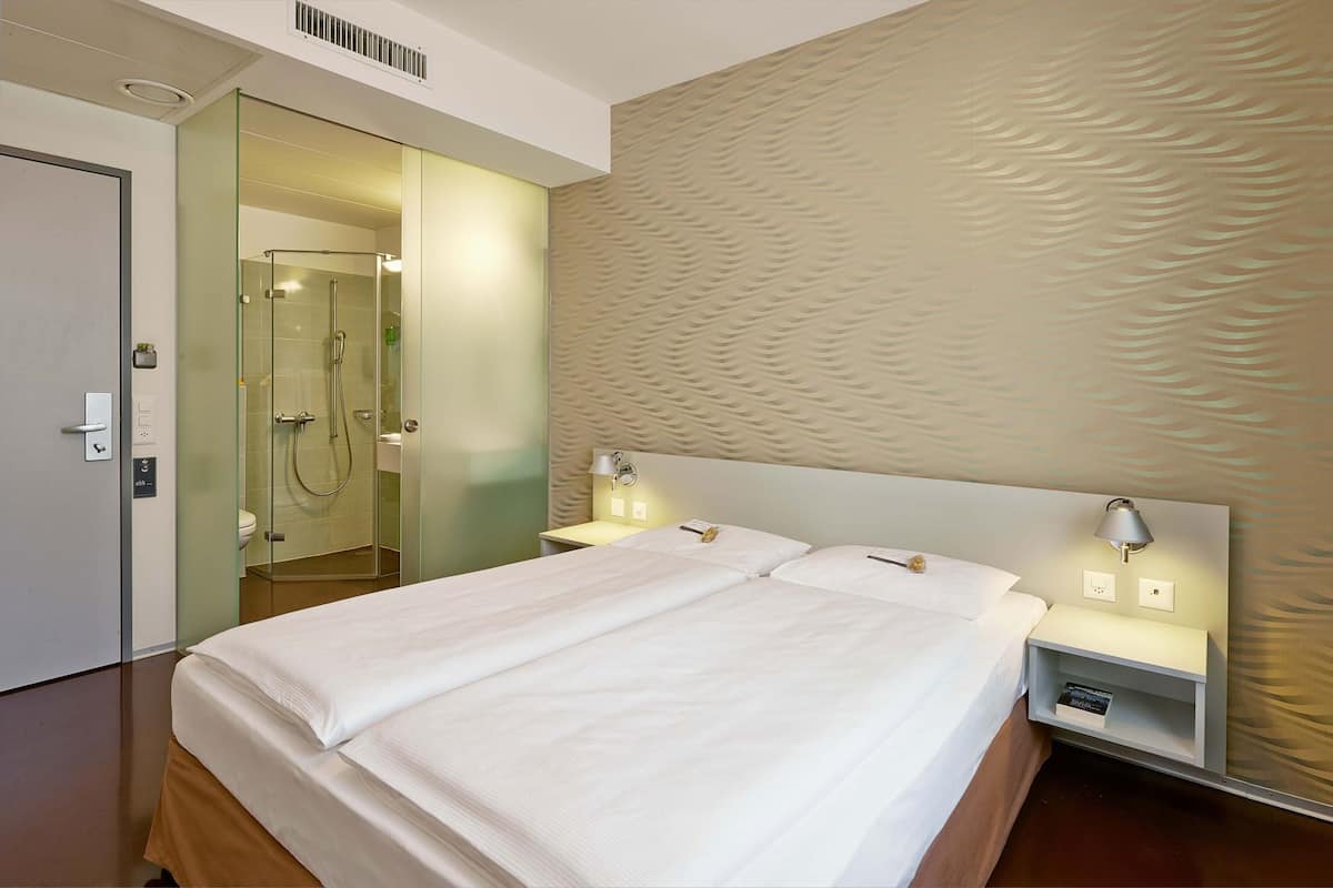 Habitación de hotel con una cama grande, dos mesitas de noche con lámparas, cabina de ducha de vidrio y una pared con textura.