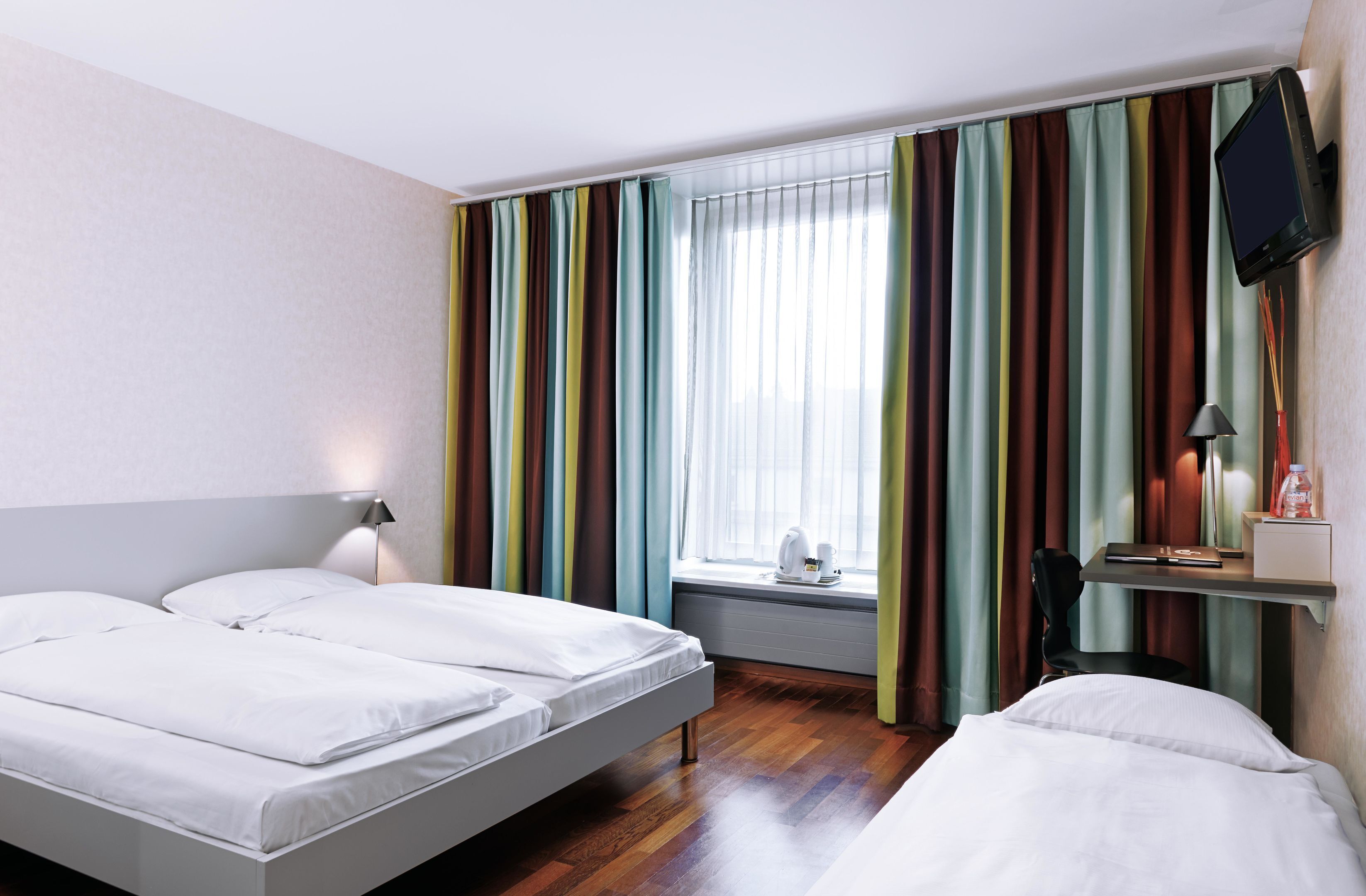 A hotel room with two beds, a TV mounted on the wall, a window with curtains, and a bedside table with a lamp.
