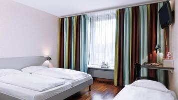 A hotel room with two beds, a TV mounted on the wall, a window with curtains, and a bedside table with a lamp.