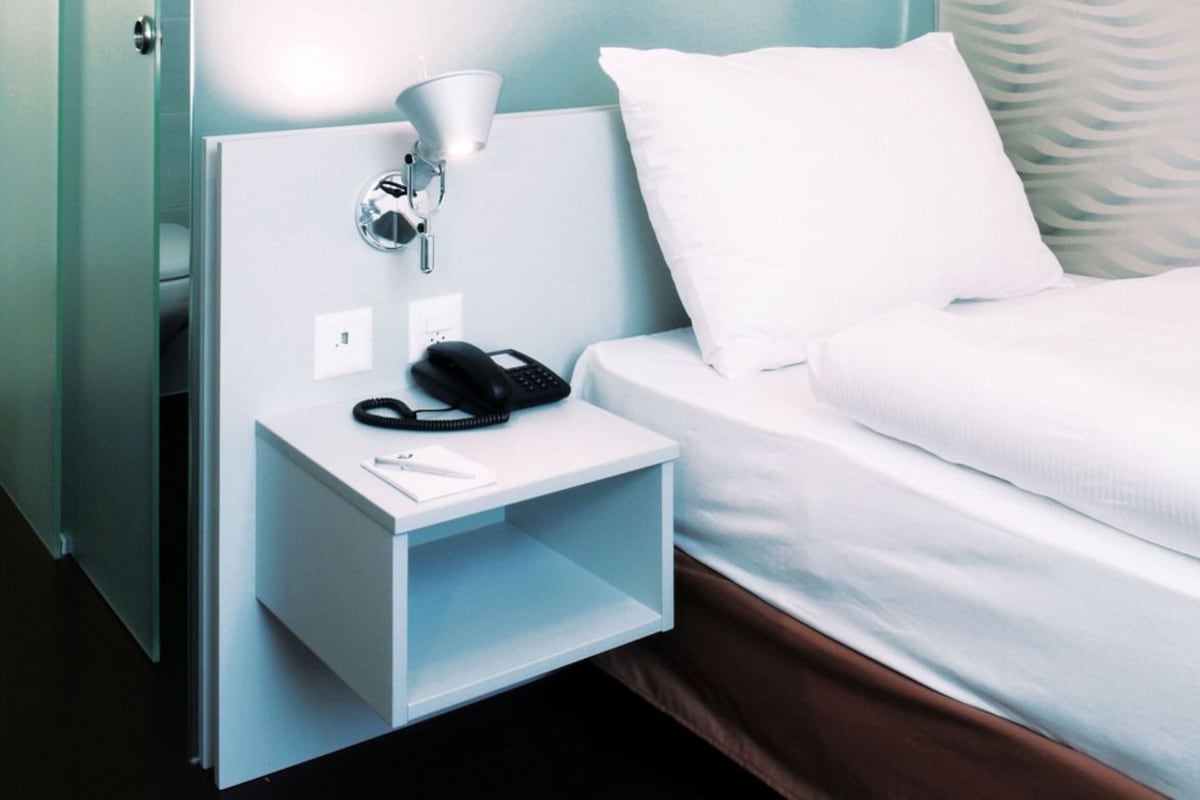 Quarto de hotel com cama, mesa de cabeceira com luminária e telefone, e parede com padrão de folhas.