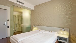 Una habitación de hotel moderna con una cama grande, lámparas de noche, una mesita de noche y una zona de ducha con paredes de vidrio.