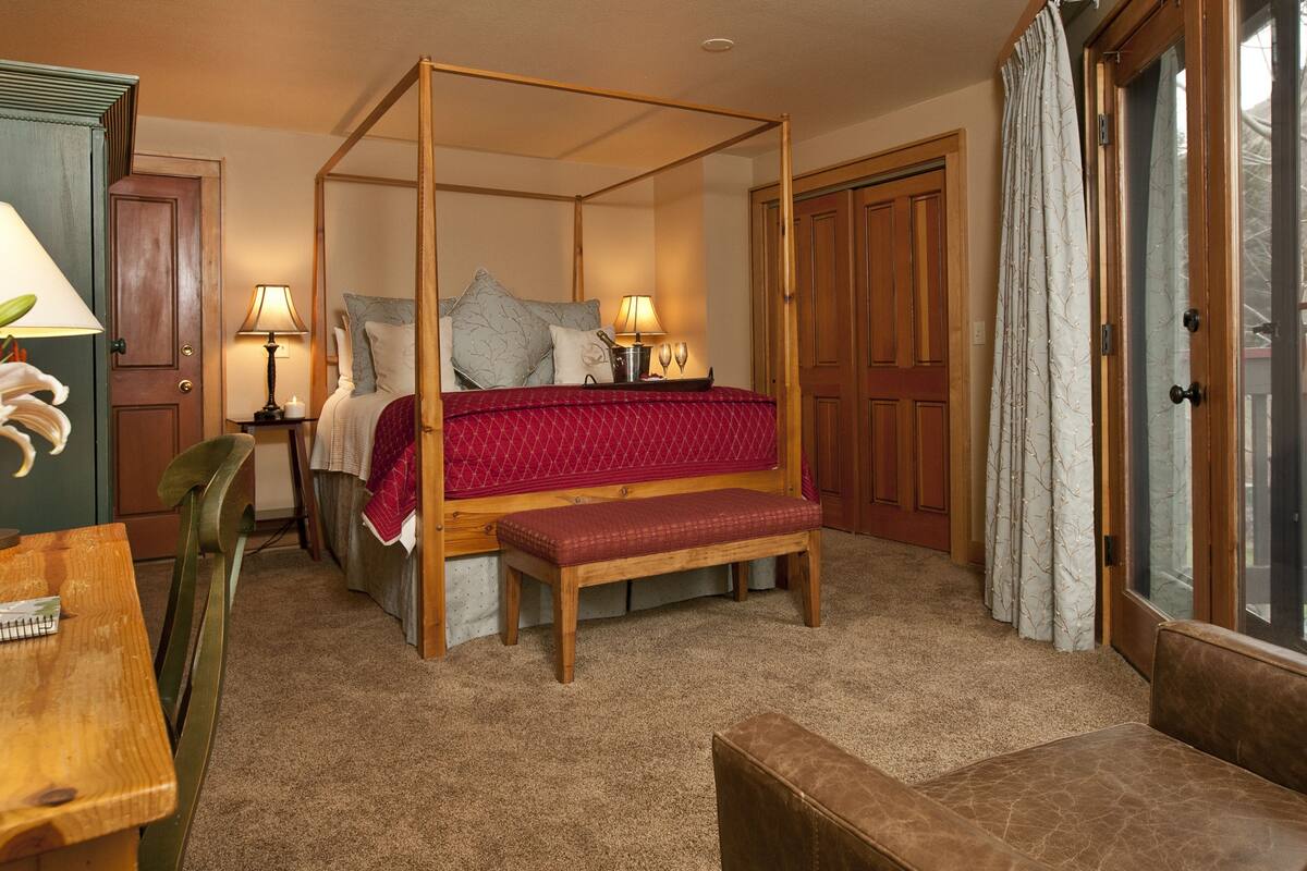 A four-poster bed with a red bedspread, two bedside lamps, a wooden bench, a wooden desk with a chair, and a door with a glass panel.