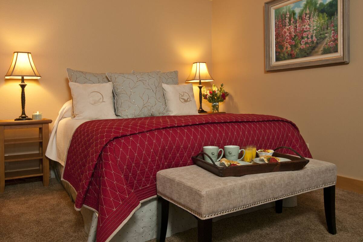 A neatly made bed with a red quilt, two bedside lamps, a tray with breakfast items, and a painting on the wall.