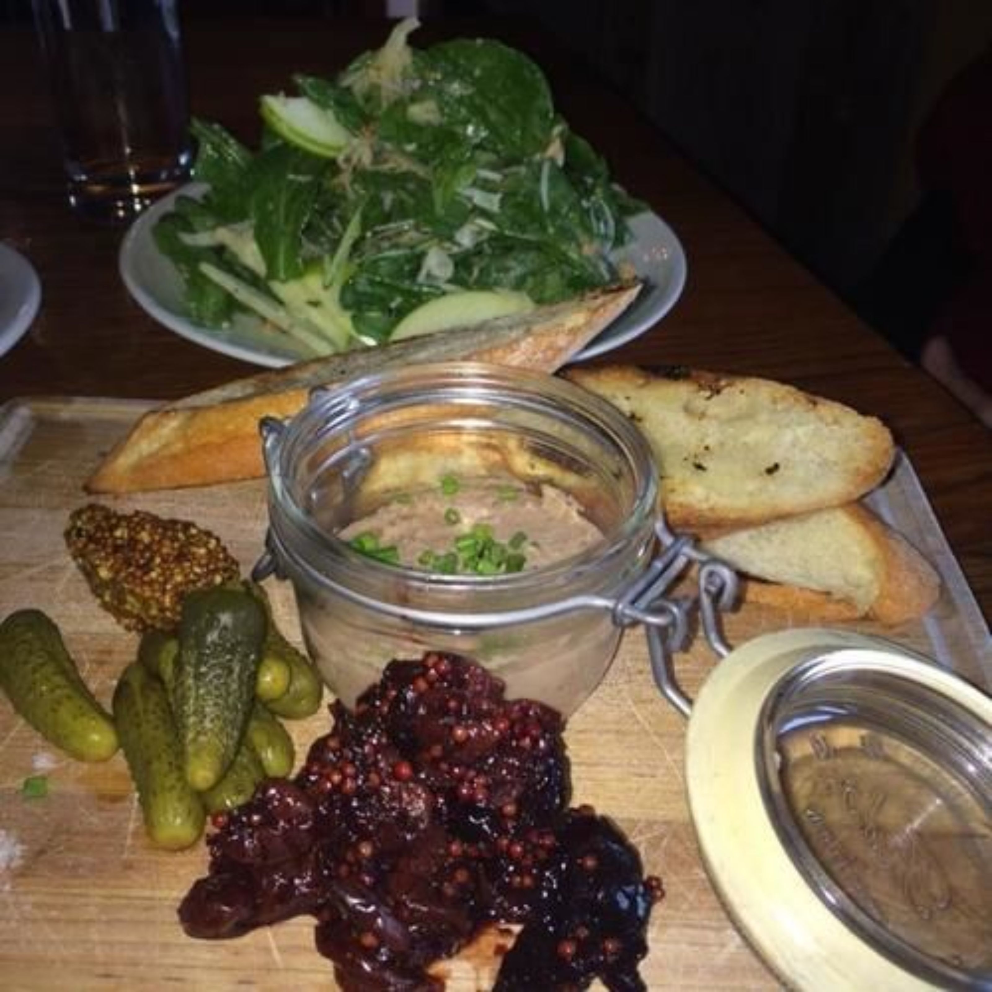 A wooden table with a glass jar of spread, pickles, and bread.