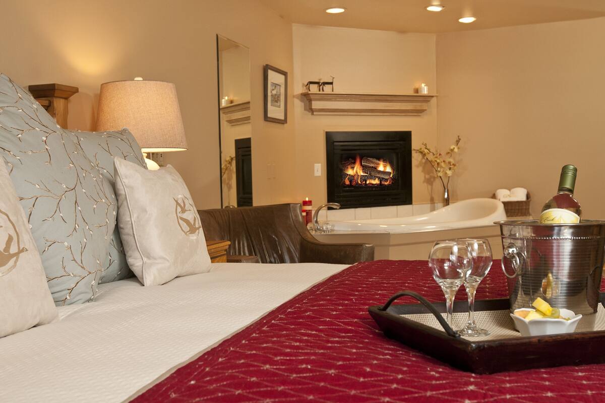 A hotel room with a bed, a fireplace, a bathtub, and a tray with glasses and a bottle.