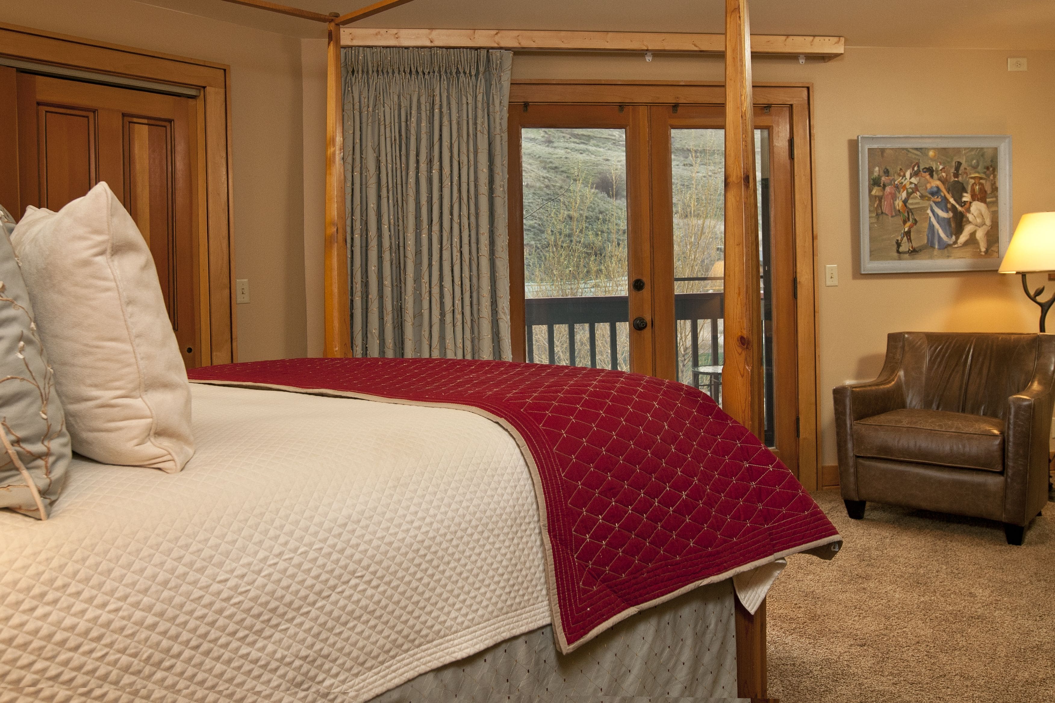 A bedroom with a large bed, a wooden door, a view through a glass door, a chair, and a painting on the wall.