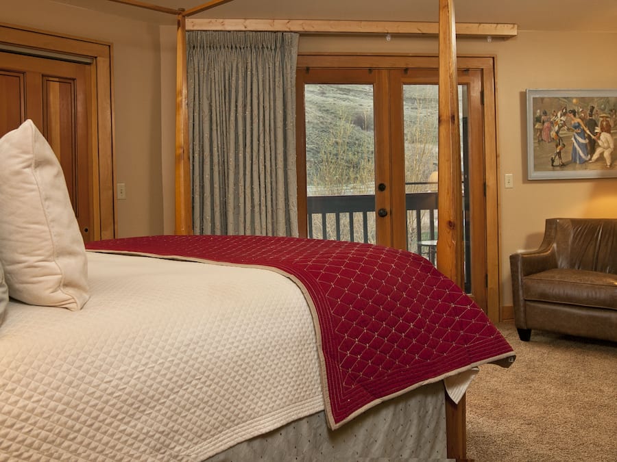 A bedroom with a large bed, a wooden door, a view through a glass door, a chair, and a painting on the wall.