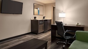 A hotel room with a flat-screen TV mounted on the wall, a kitchenette with a microwave and refrigerator, a desk with a chair, and a green upholstered chair.