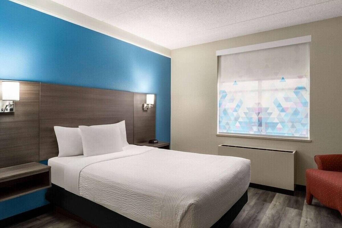 A hotel room with a bed, bedside tables, a chair, a window with a geometric pattern, and a wall with a blue and beige color scheme.