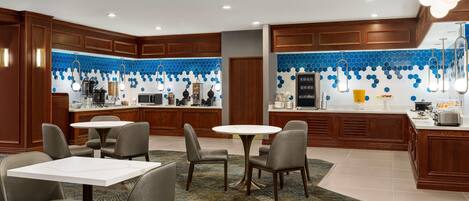 A modern kitchen with a blue hexagonal backsplash, wooden cabinets, and a central island with a sink and stove. There are two round tables with chairs in the dining area.