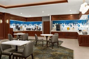 A modern kitchen with a blue hexagonal backsplash, wooden cabinets, and a central island with a sink and stove. There are two round tables with chairs in the dining area. - La Quinta Inn & Suites by Wyndham Madison American Center (Madison)
