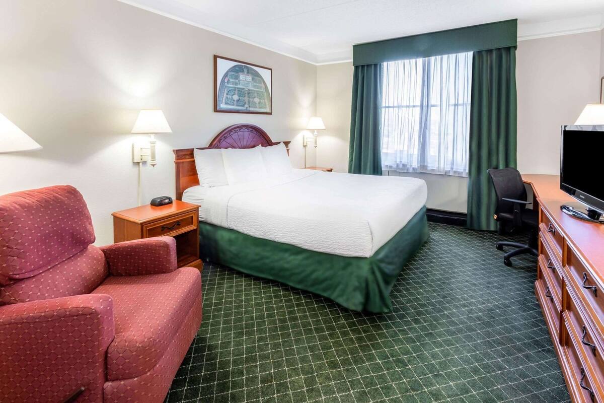 A hotel room with a bed, a desk with a television, a chair, a nightstand, and a window with curtains.