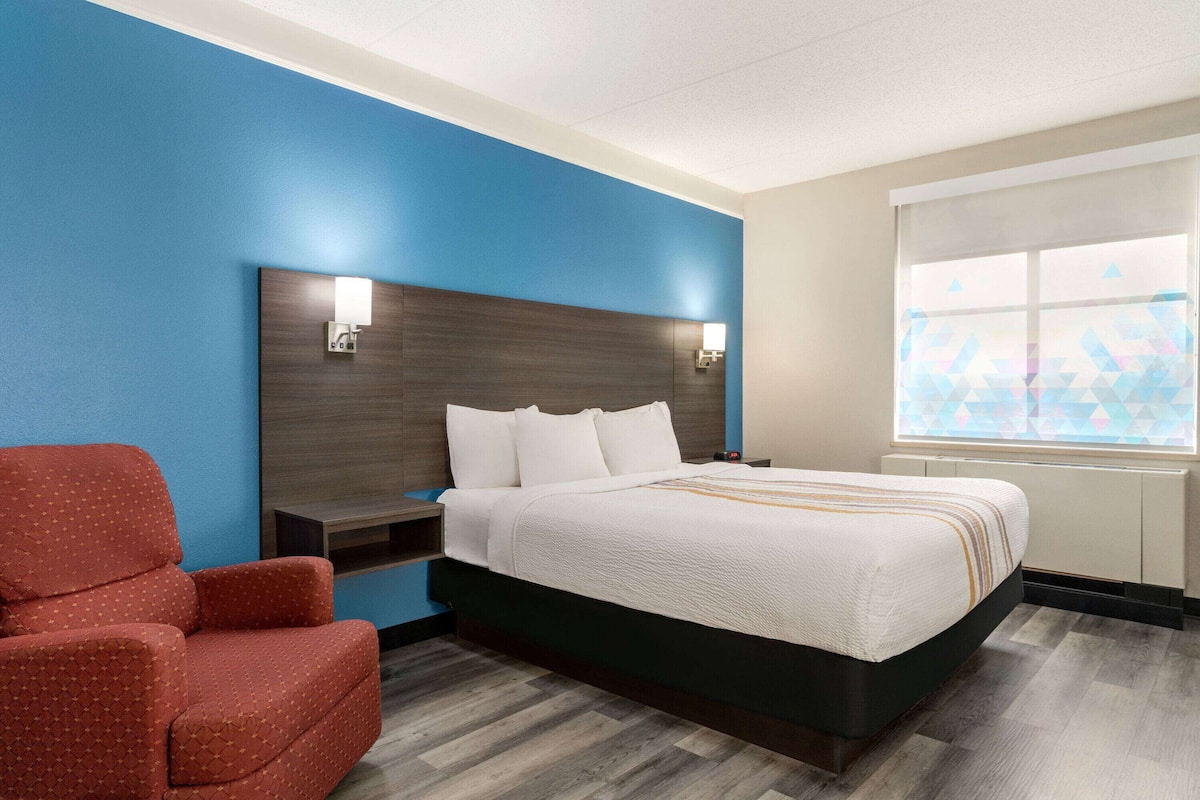 A hotel room with a bed, a red armchair, a nightstand, and a window with geometric patterned blinds.