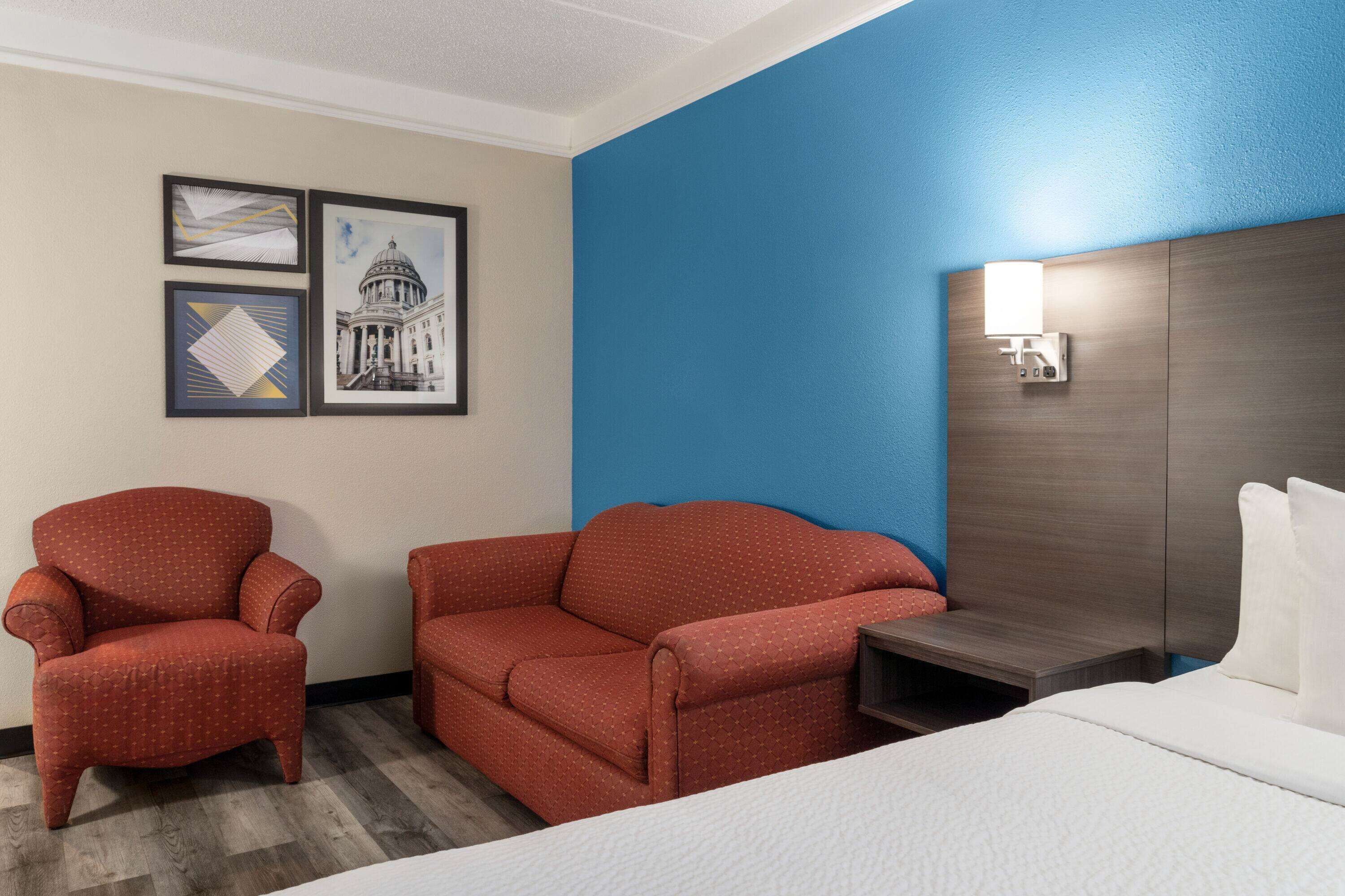A hotel room with a bed, a red armchair, a red sofa, a nightstand, and a wall-mounted lamp.