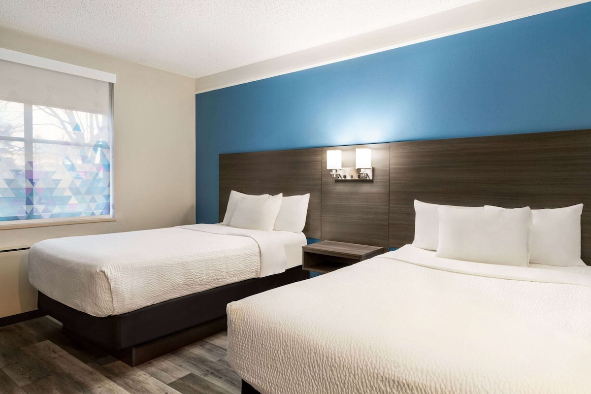 A hotel room with two beds, a large window with geometric blinds, and a dark wood headboard with wall-mounted lights.