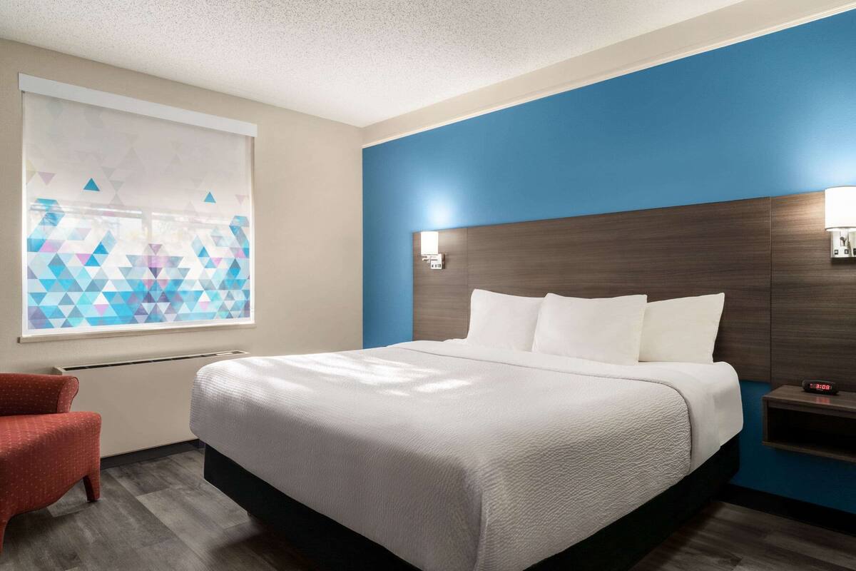 A hotel room with a large bed, a red armchair, a window with geometric blinds, and a nightstand with a lamp.
