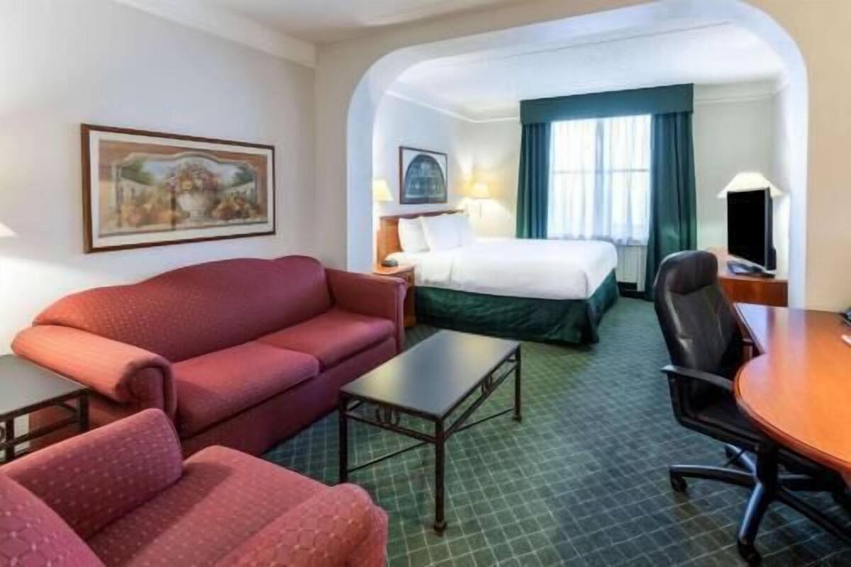 A hotel room with a red sofa, a bed with green bedding, a desk with a chair, and a television.