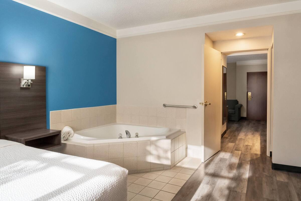 A hotel room with a large bathtub, a wooden nightstand, a bed with a white textured bedspread, and a blue accent wall.