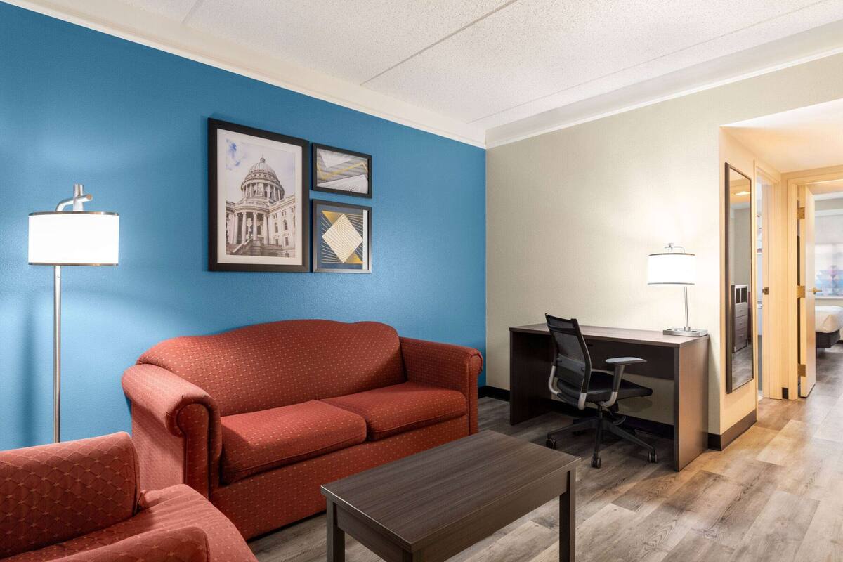 A hotel room with a blue accent wall, a red sofa, a desk with a chair, and a small table.