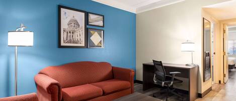 A hotel room with a blue accent wall, a red sofa, a desk with a chair, and a small table.