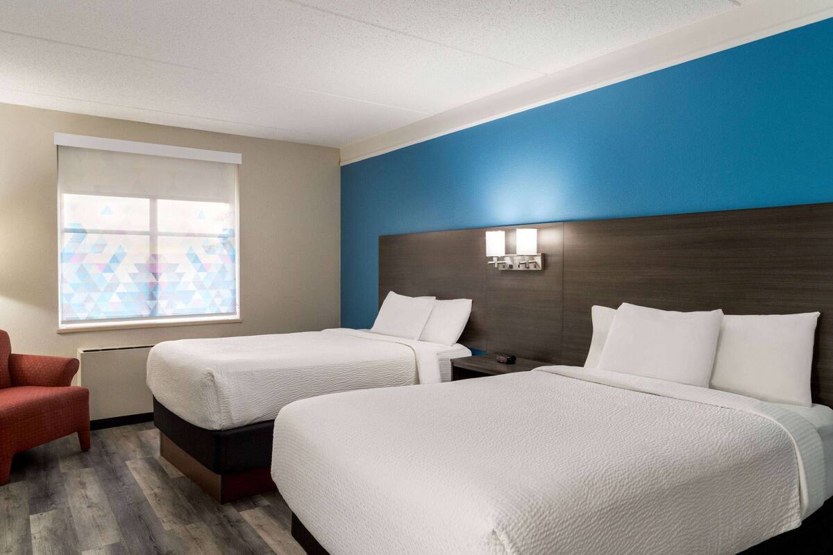 A hotel room with two beds, a red chair, a window with a geometric pattern, and a blue accent wall.
