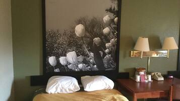 Una habitación de hotel con cama, almohadas, un escritorio con lámpara y un cuadro en la pared.