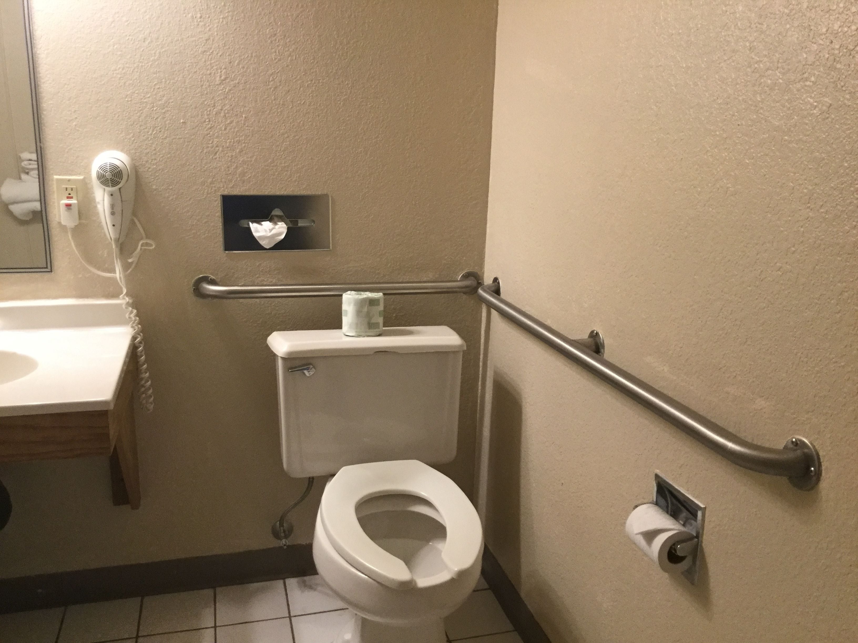 A bathroom with a toilet, a hand dryer, a paper towel dispenser, and a handrail.