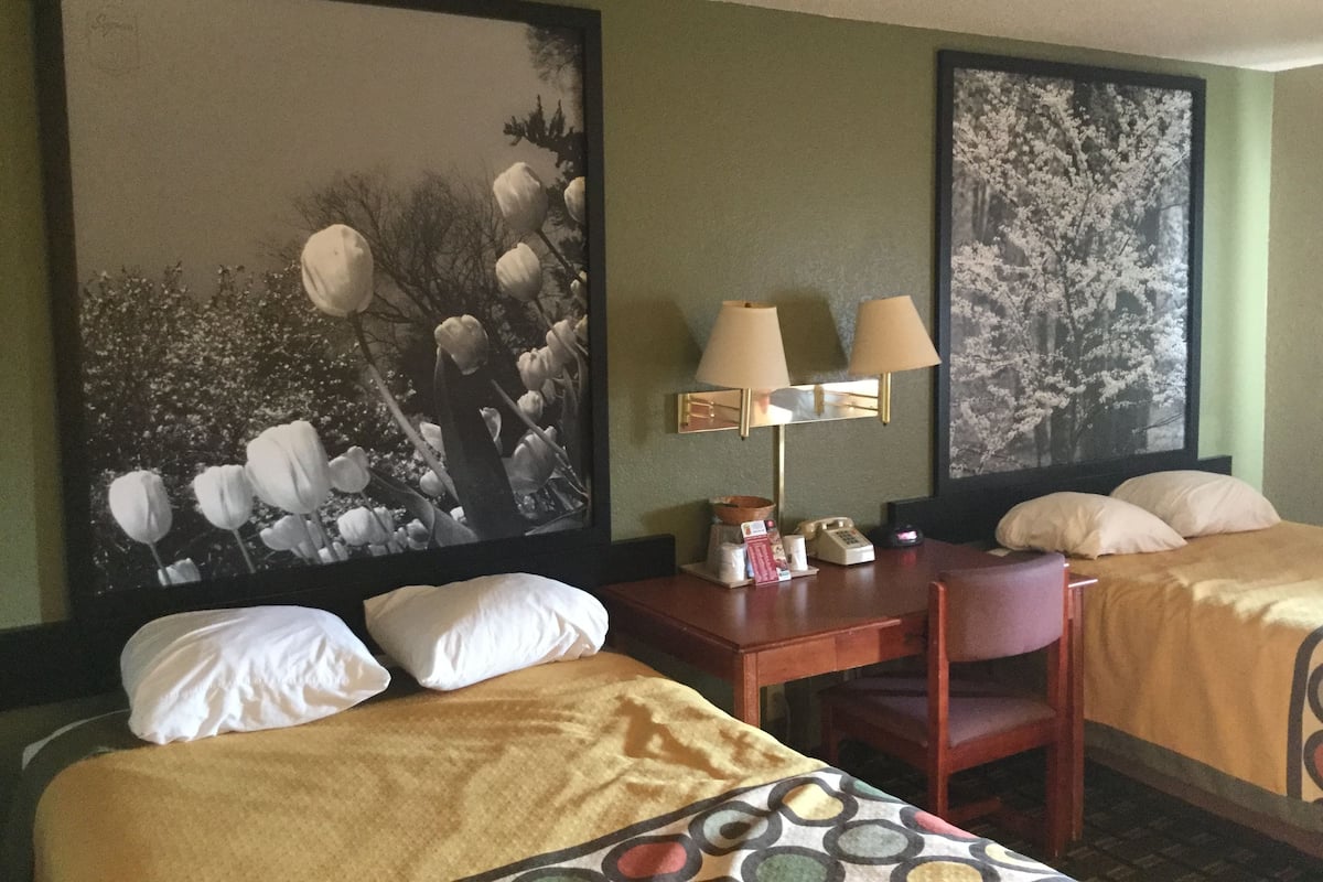 A hotel room with two beds, a desk with a lamp, and two paintings on the wall.