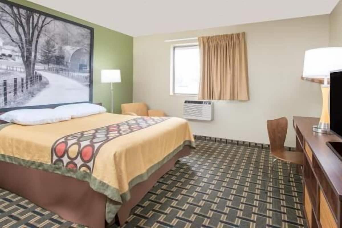 A hotel room with a bed, a desk, a chair, a window with curtains, an air conditioning unit, and a wall-mounted TV.