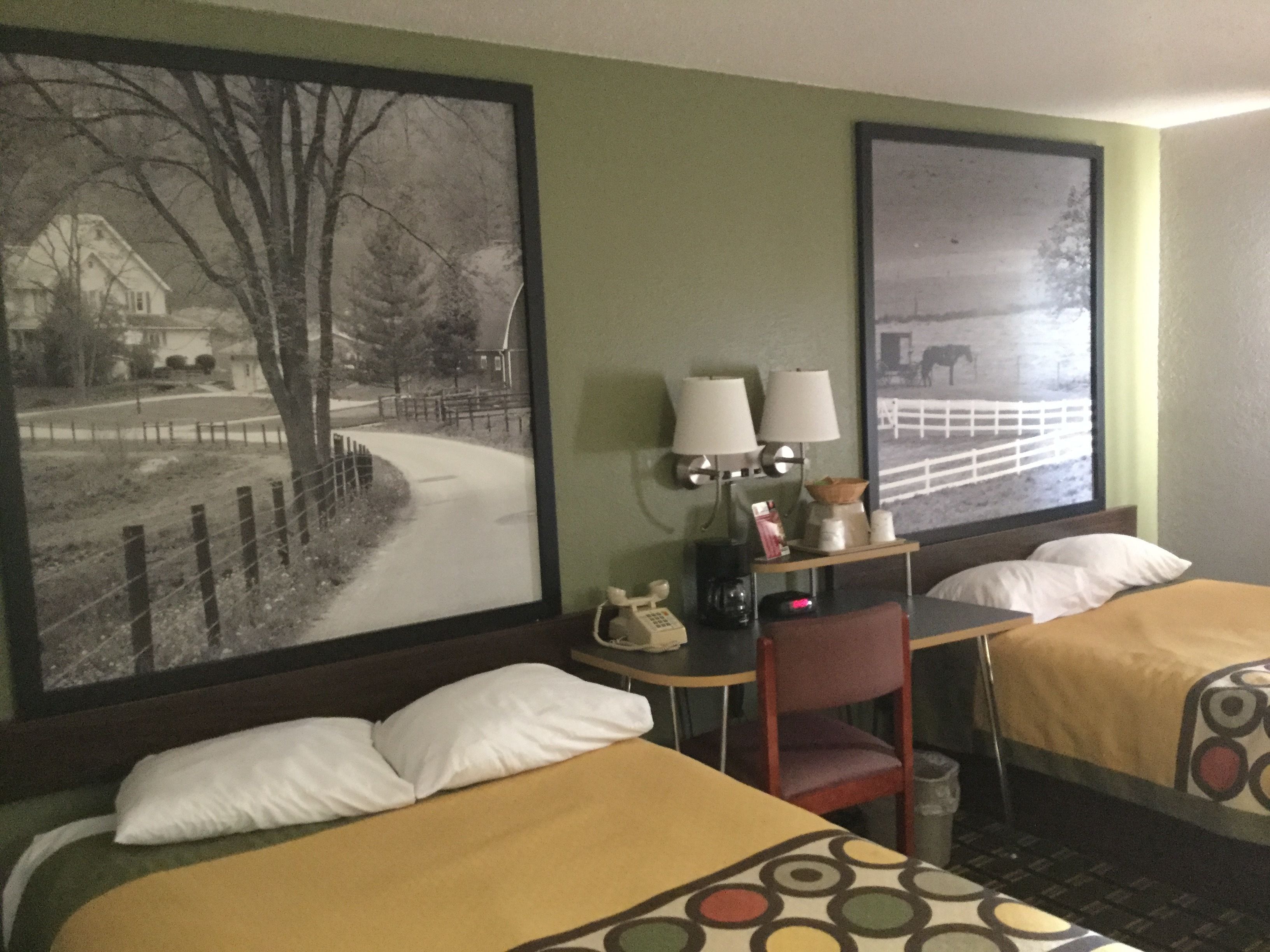Une chambre d’hôtel avec deux lits, un bureau avec une lampe, un téléphone et un tableau représentant un paysage rural accroché au mur.