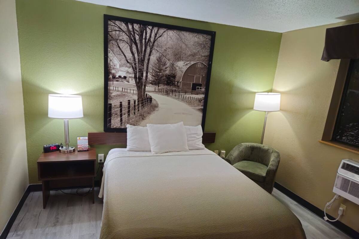 A hotel room with a bed, two bedside lamps, a chair, a nightstand with a clock, and a large framed picture on the wall.