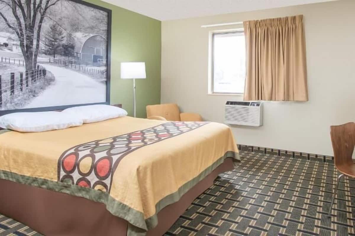 A hotel room with a bed, a desk, a chair, a lamp, a window with curtains, a wall-mounted air conditioner, and a large painting on the wall.